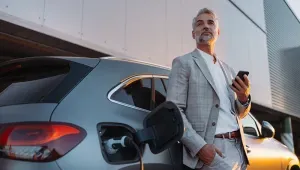 man van middelbare leeftijd met een smartphone in de hand leunt tegen een opladende elektrische auto