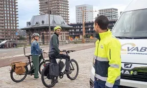 Wegenwachter staat bij een VAB‑voertuig terwijl twee personen met hun fietsen stoppen op een plein
