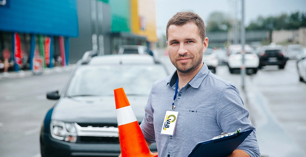 VAB-rijlesgever staat voor een auto met een clipboard en oranje kegeltje in de hand