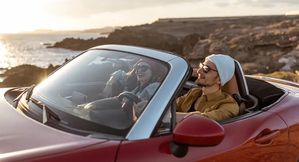 man en vrouw zitten in een rode cabrio aan de rand van een klif bij ondergaande zon boven de zee