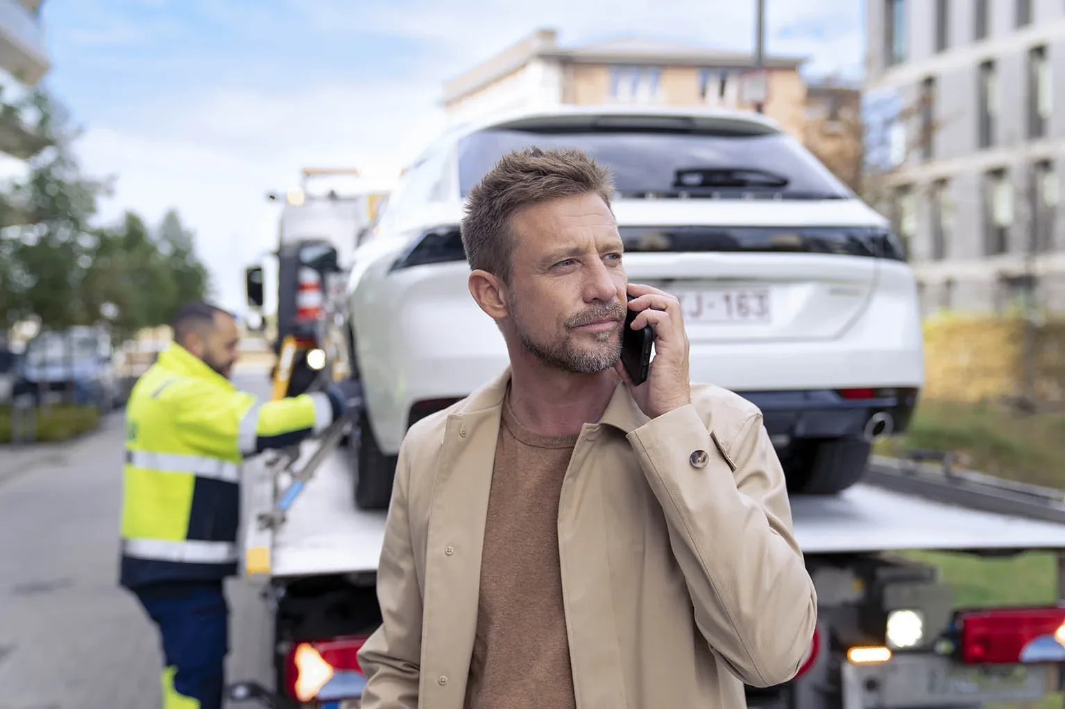 man van middelbare leeftijd telefoneert terwijl hij voor een takelwagen staat waarop zijn auto bevestigd wordt door een VAB-wegenwachter