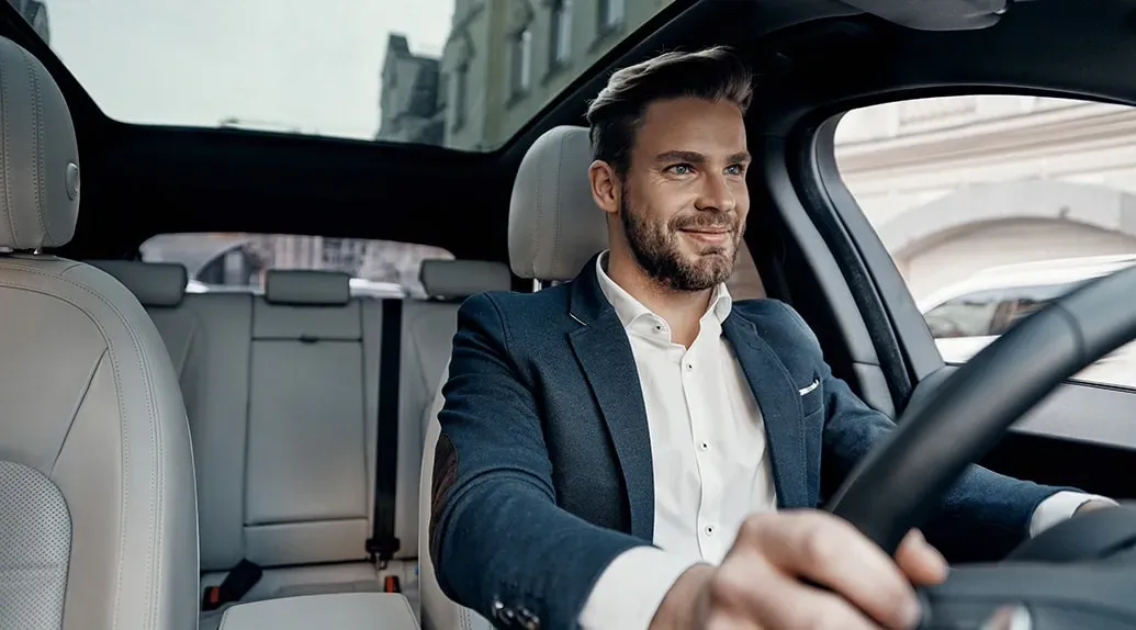 een man met korte baard, kostuum en hemd zit achter het stuur van een auto met een lichtgrijs interieur