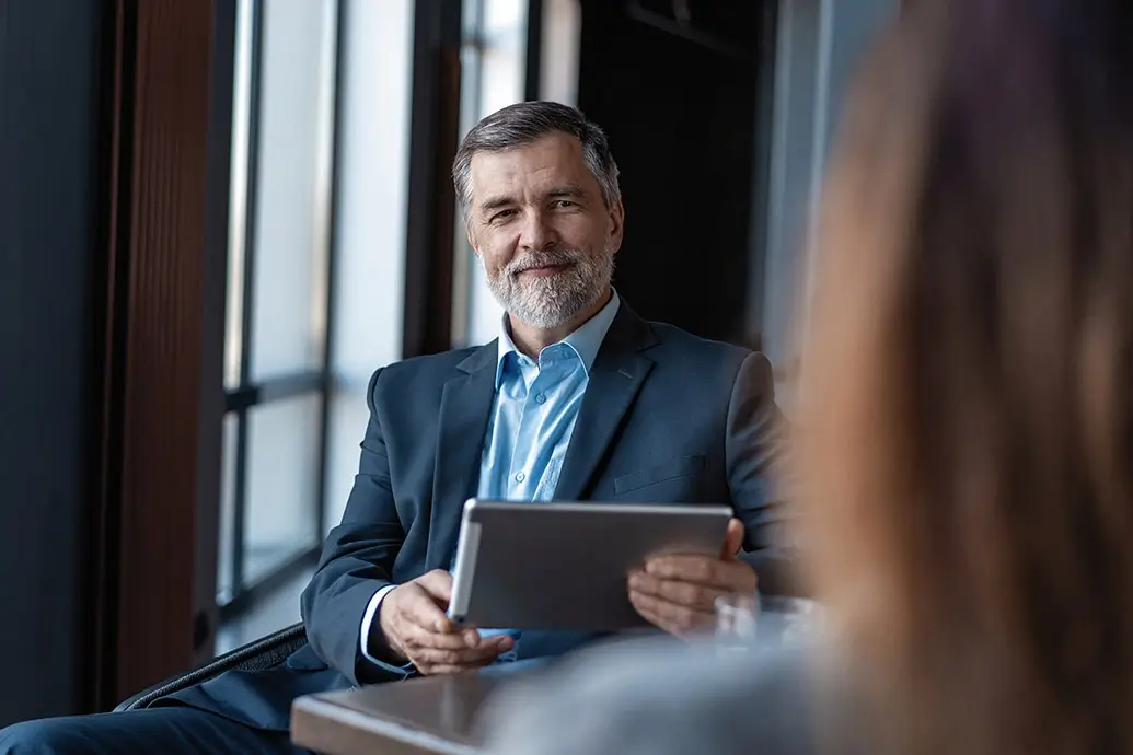 een man met grijze baard, maatpak en hemd houdt een tablet vast terwijl hij een roodharige vrouw interviewt