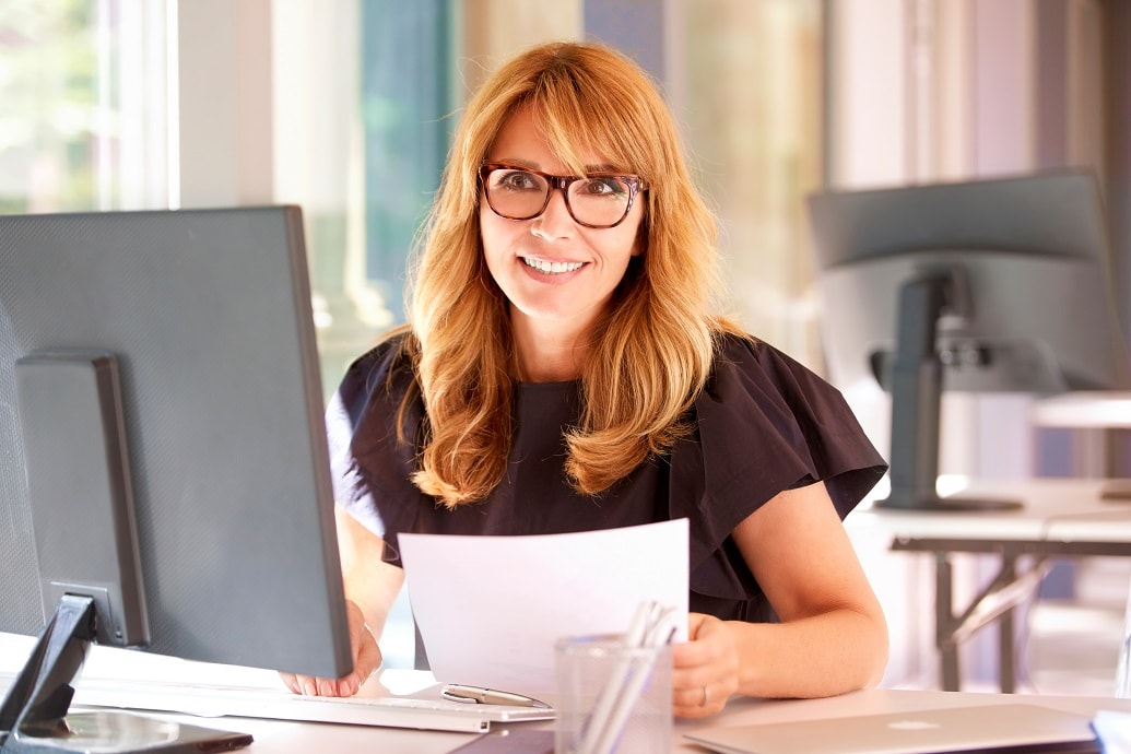 vrouw met glimlach en bril zit aan een computerscherm met papieren in de hand