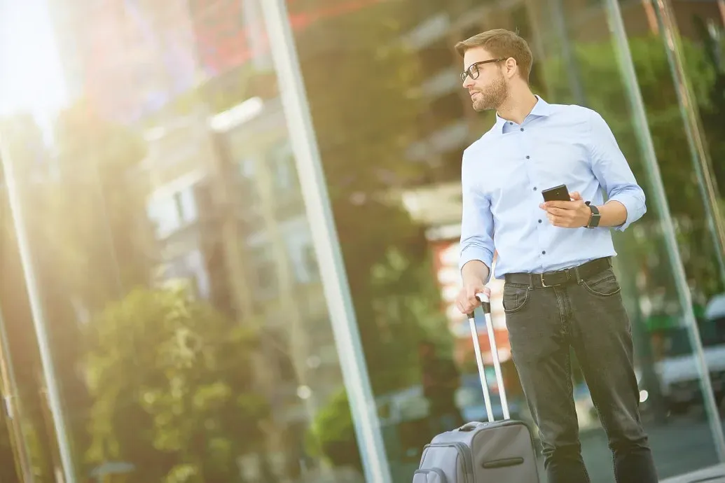man in zakelijke kledij staat voor een glazen gevelwand met een koffertje in 1 hand en gsm in de andere hand