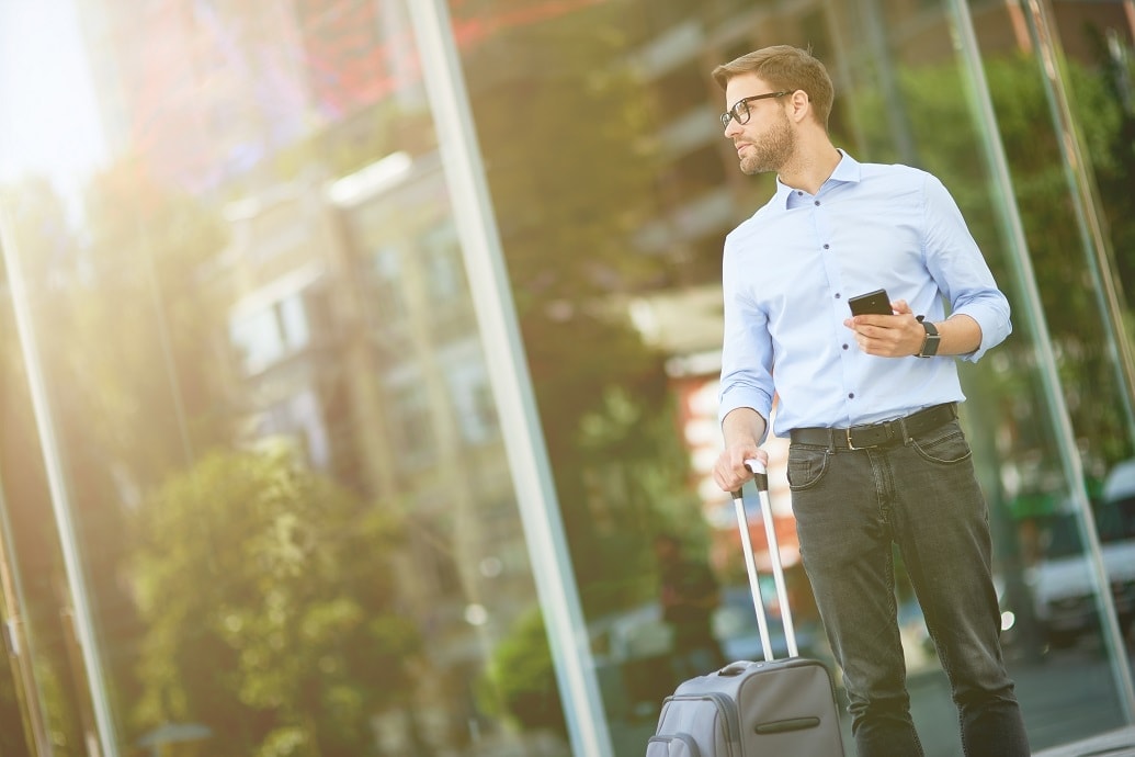 man in zakelijke kledij staat voor een glazen gevelwand met een koffertje in 1 hand en gsm in de andere hand