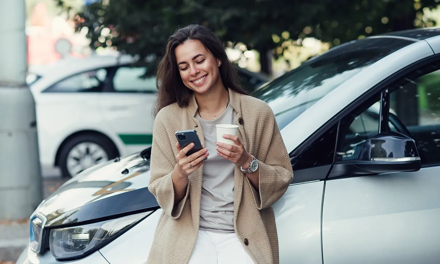 Vrouw kijkt naar telefoon met koffie in haar hand naast een auto