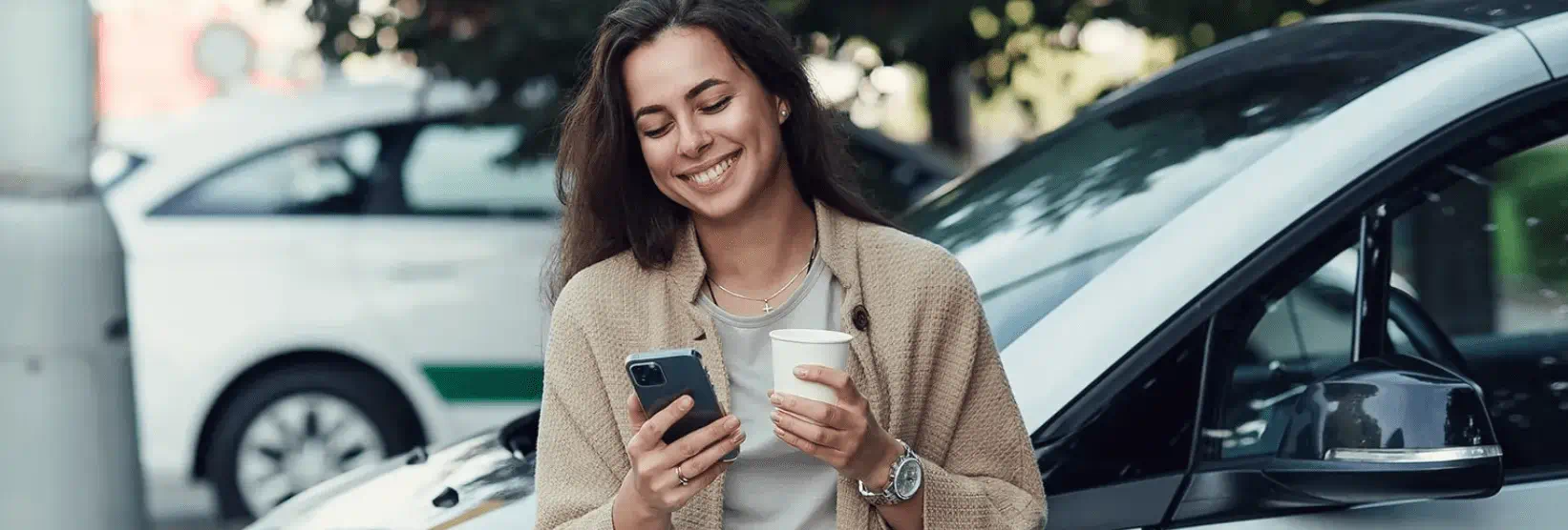 Vrouw kijkt naar telefoon met koffie in haar hand naast een auto