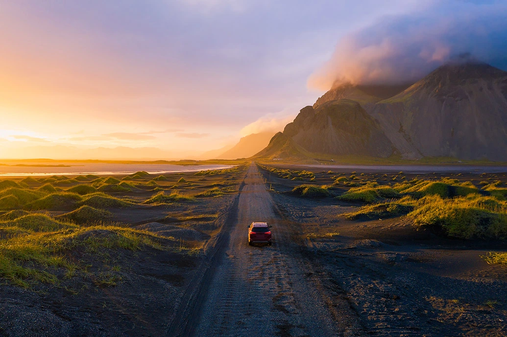 een rode auto rijdt op een landweg met links een zonsondergang en rechts een bergkam