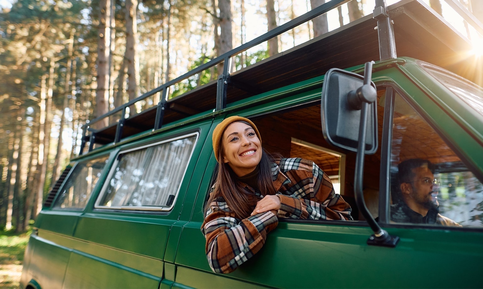 Vrouw in een kampeerbusje leunt uit het raam terwijl ze met haar partner in een bosrijk landschap geparkeerd staat.