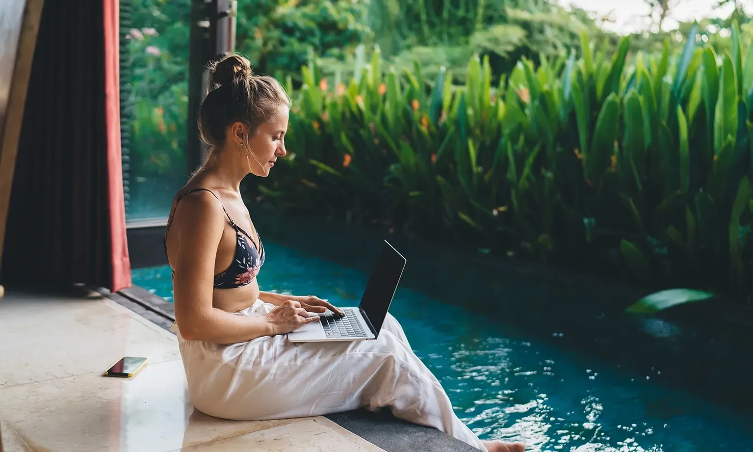 Personne travaille sur un ordinateur près d’une piscine entourée de plantes tropicales.