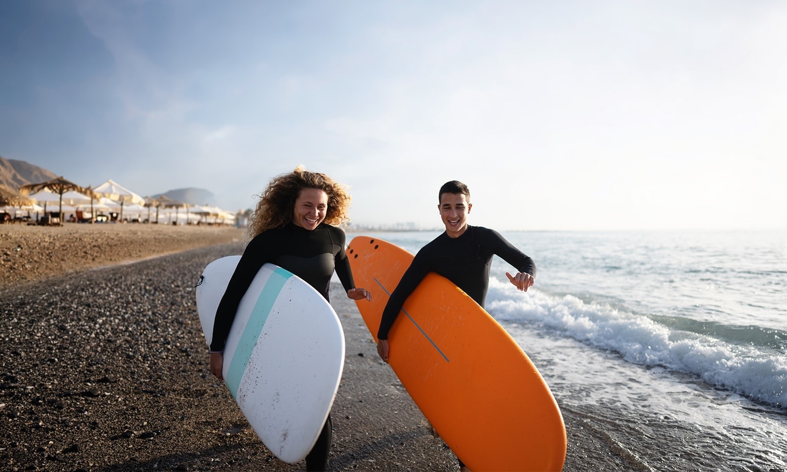 Twee personen lopen met surfboards langs de waterlijn op een zonnig strand.