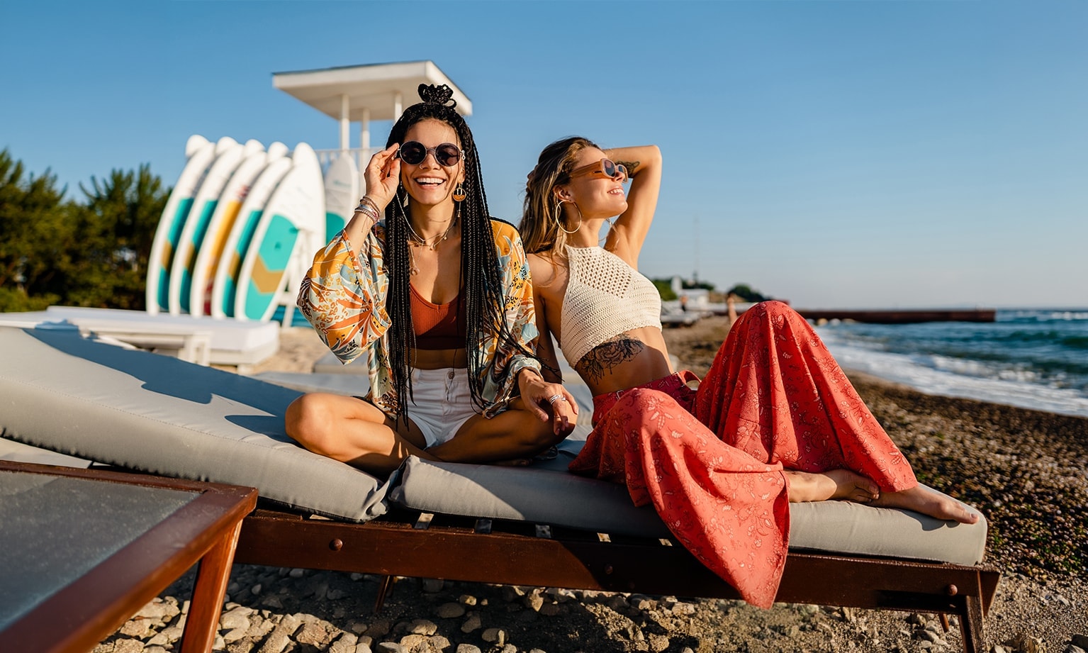 Twee personen ontspannen op strandligbedden bij zee met surfboards op de achtergrond.