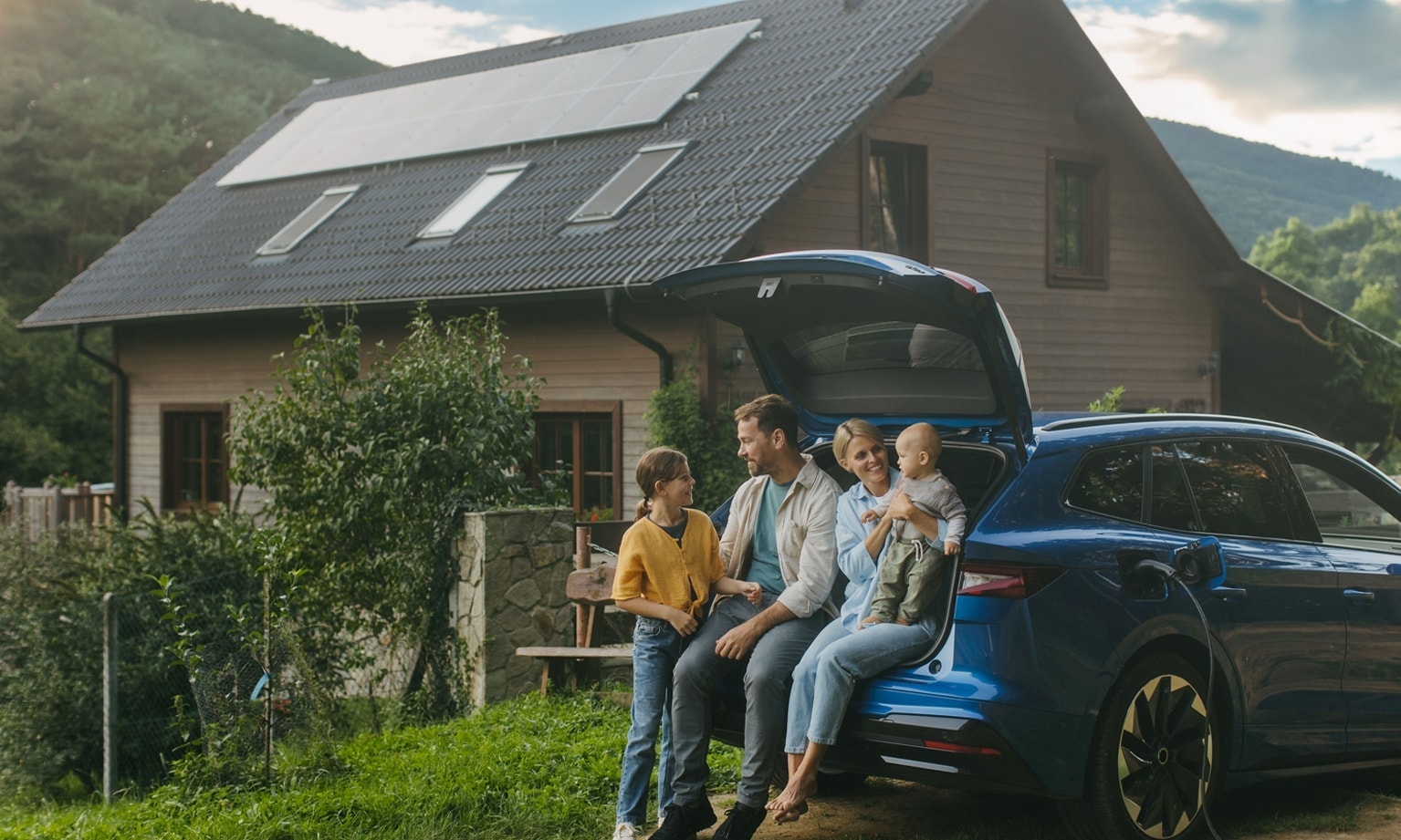 Famille assise près d’un coffre de voiture ouvert devant une maison avec panneaux solaires.